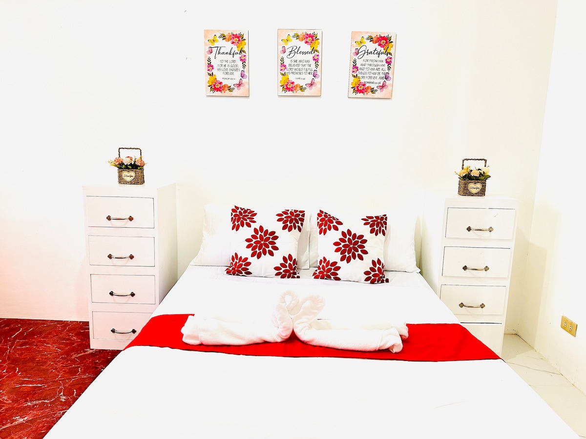A double size bed is featured, adorned with a red and white color scheme. The bed is accompanied by decorative pillows with floral patterns. Two nightstands with handles are situated on either side, and three framed floral artworks hang on the wall above.