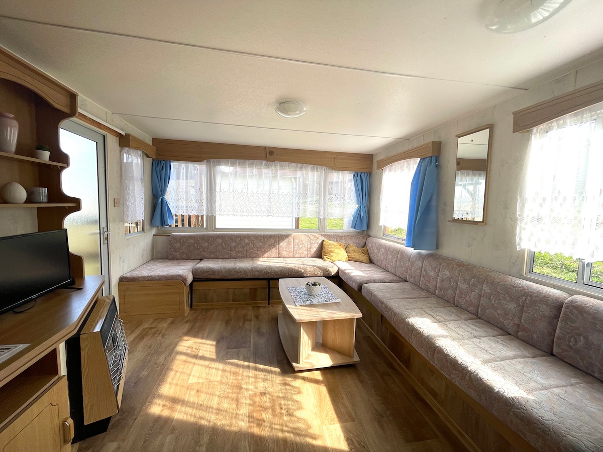 A sunlit living area features a spacious L-shaped seating arrangement with soft cushions, complemented by a central coffee table. Large windows with sheer curtains allow natural light to filter in, while light wood flooring enhances the open atmosphere.