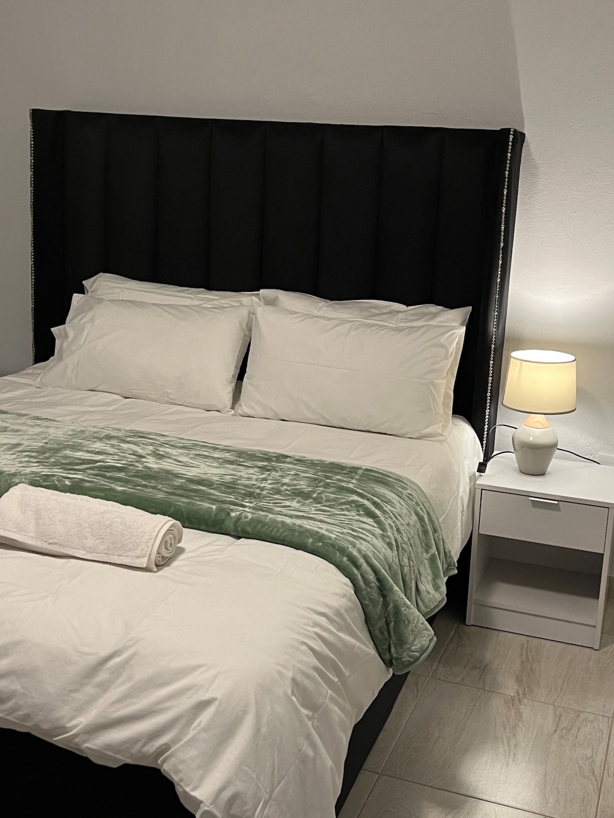 A spacious queen-size bed is dressed in crisp white linens, complemented by a soft green throw. A pair of decorative pillows adds a touch of elegance. A bedside table with a lamp provides warm lighting, enhancing the cozy atmosphere of the room.