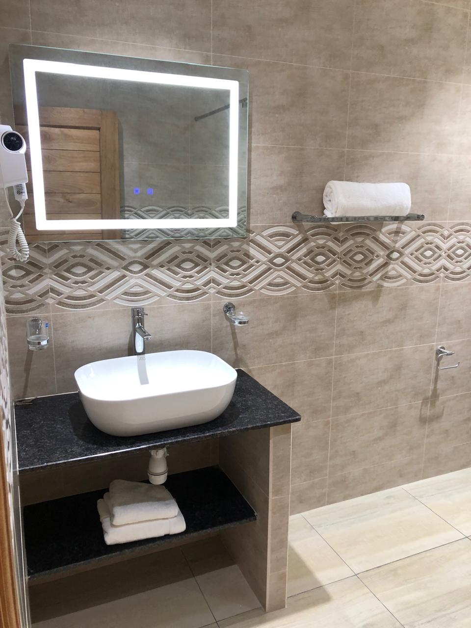 A modern bathroom features a sleek, white basin mounted on a dark countertop. A large illuminated mirror reflects the tasteful tile work and patterned accent strip. Bath towels are neatly stored on a shelf above the basin, contributing to the functional design.