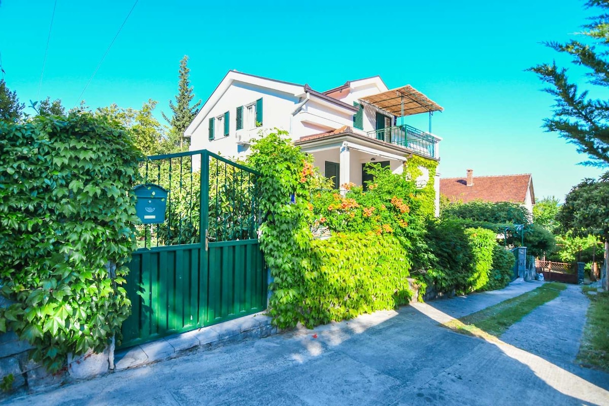 A well-maintained exterior view of a two-story house surrounded by lush greenery. The entrance features a green gate leading to a pathway, with vibrant plants climbing the walls. A balcony can be seen on the upper level, providing an open space for relaxation.