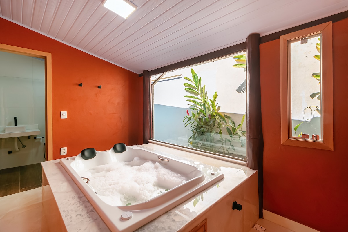 A modern bathroom features a spacious whirlpool tub positioned against a backdrop of warm red walls. Natural light filters through large windows, illuminating the surrounding greenery that peeks in from outside.