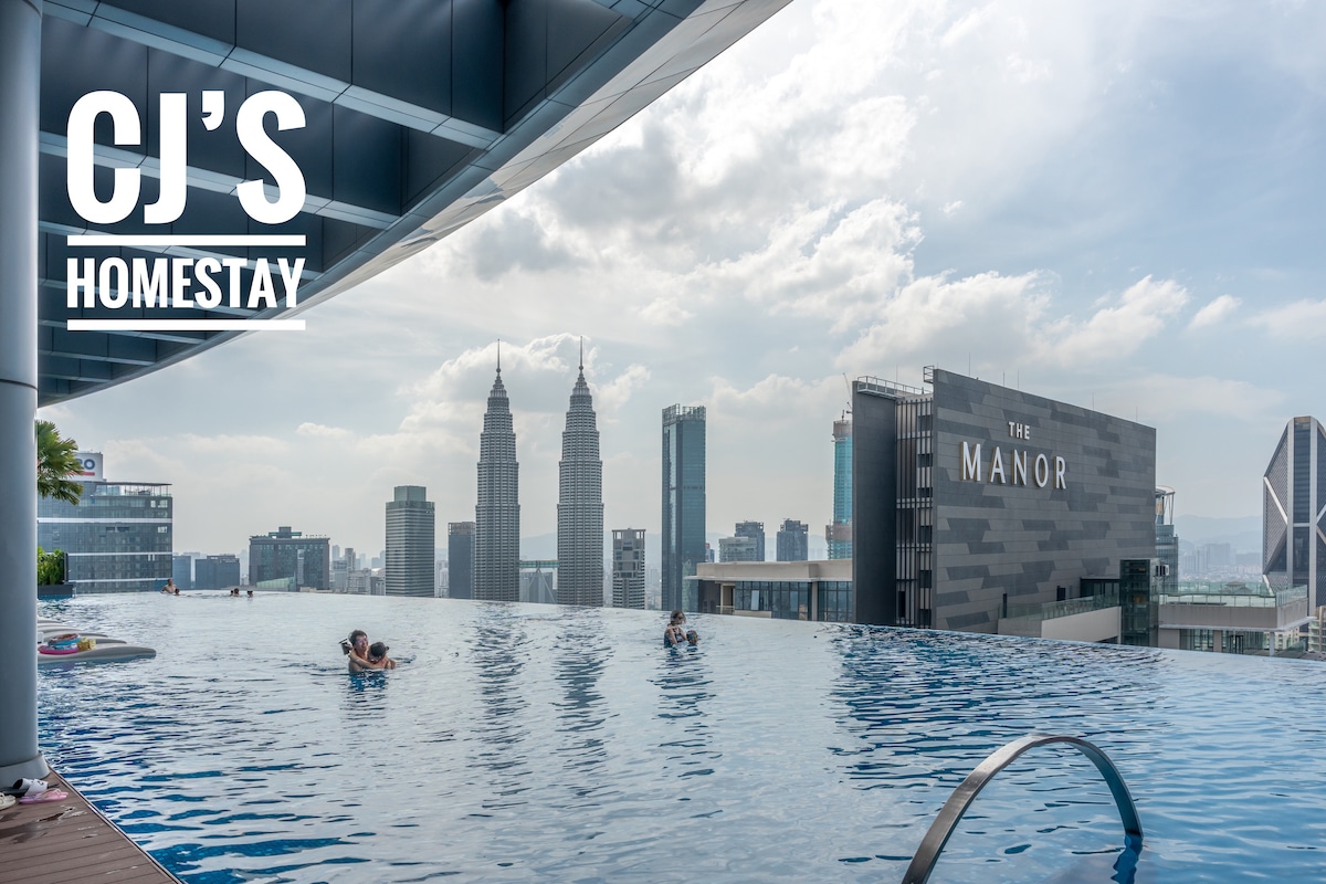 An infinity edge pool extends towards the edge, offering panoramic views of the skyline, including the iconic Petronas Twin Towers. Gentle waves are created by swimmers, while the surrounding area is characterized by modern buildings and soft cloud formations above.