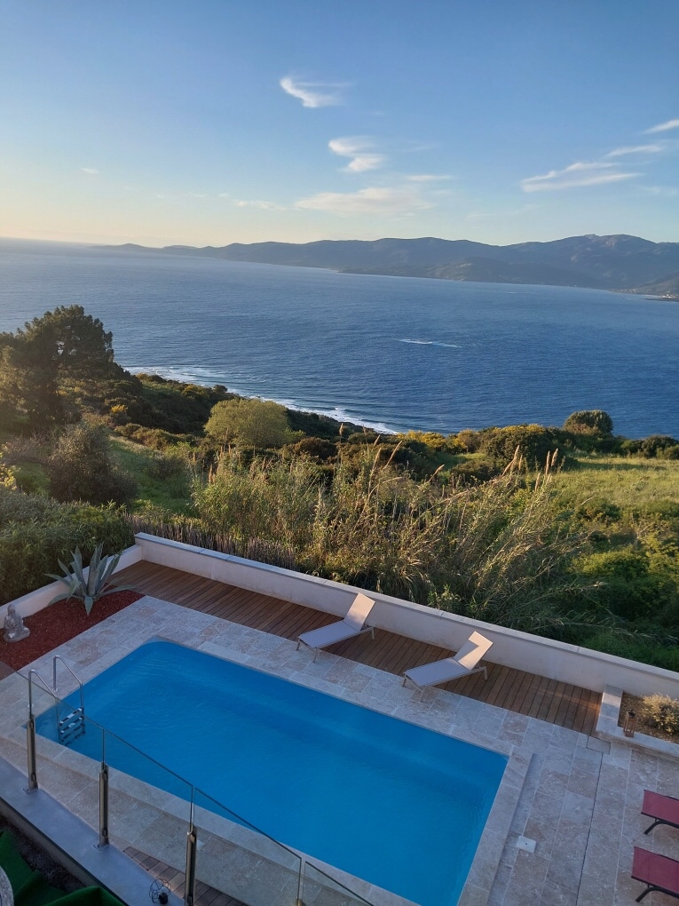 A private pool is situated on a terrace, overlooking a vast expanse of the sea and distant mountains. Sun loungers are positioned beside the pool, and lush greenery surrounds the area, adding to the natural beauty of the landscape.
