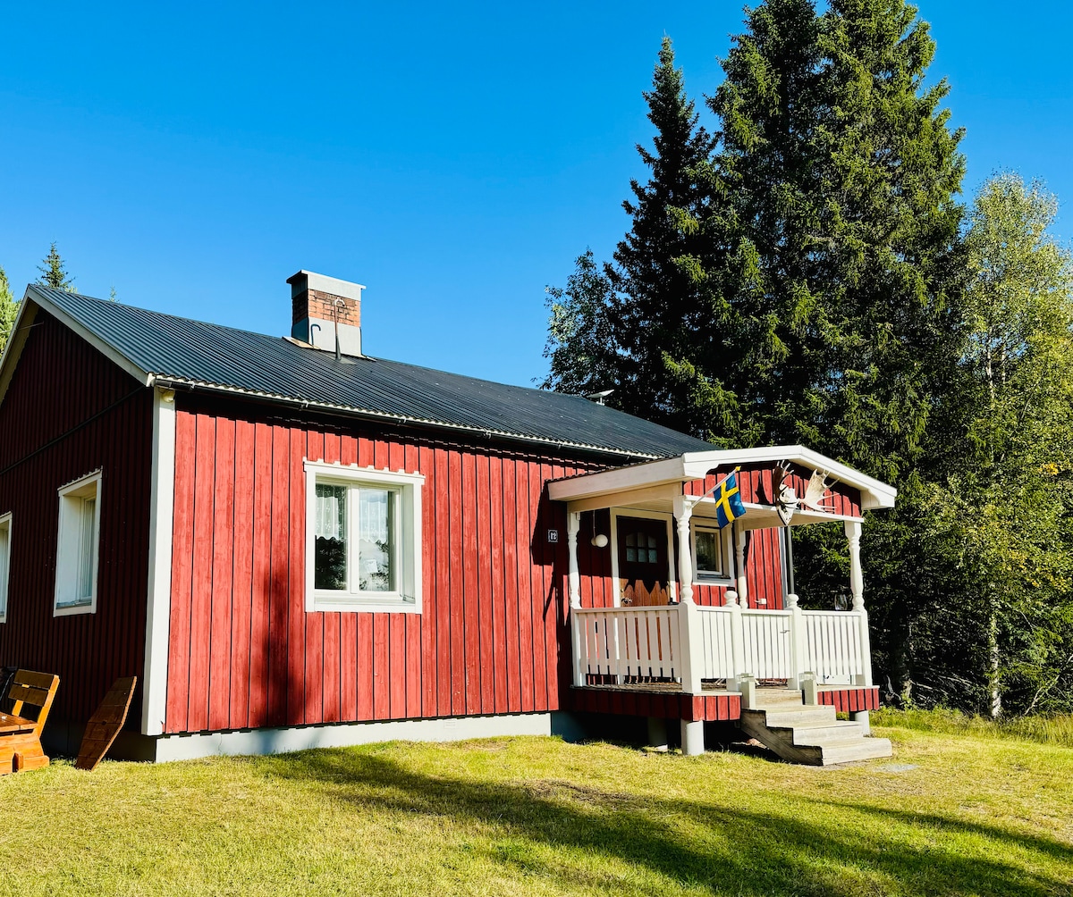 A red wooden cabin is depicted with a pitched roof and a welcoming front porch. Surrounding greenery includes tall trees and a clear blue sky. The porch features a railing with decorative elements, creating an inviting entrance to the home.