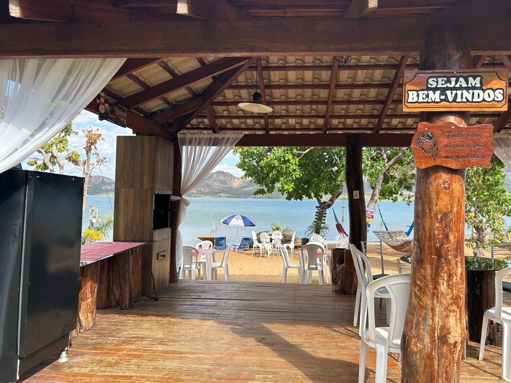 A welcoming outdoor space features wooden beams and a thatched roof. A charcoal grill is positioned on one side, while white plastic tables and chairs are arranged for seating. The serene lake is visible through the open area, enhancing the connection with nature.