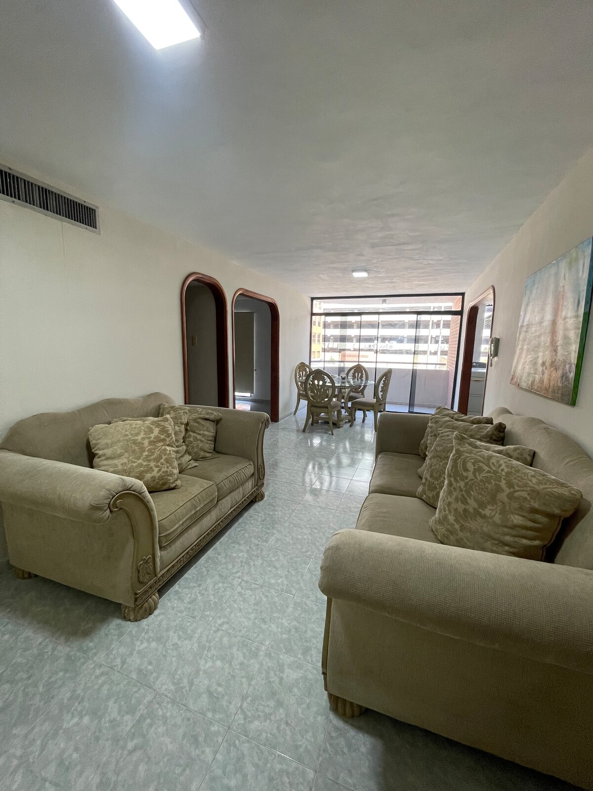 A living area is shown with two inviting sofas, adorned with decorative cushions. A dining set for six is visible in the background, surrounded by large windows that allow natural light to fill the space. The floors are tiled and reflect light.