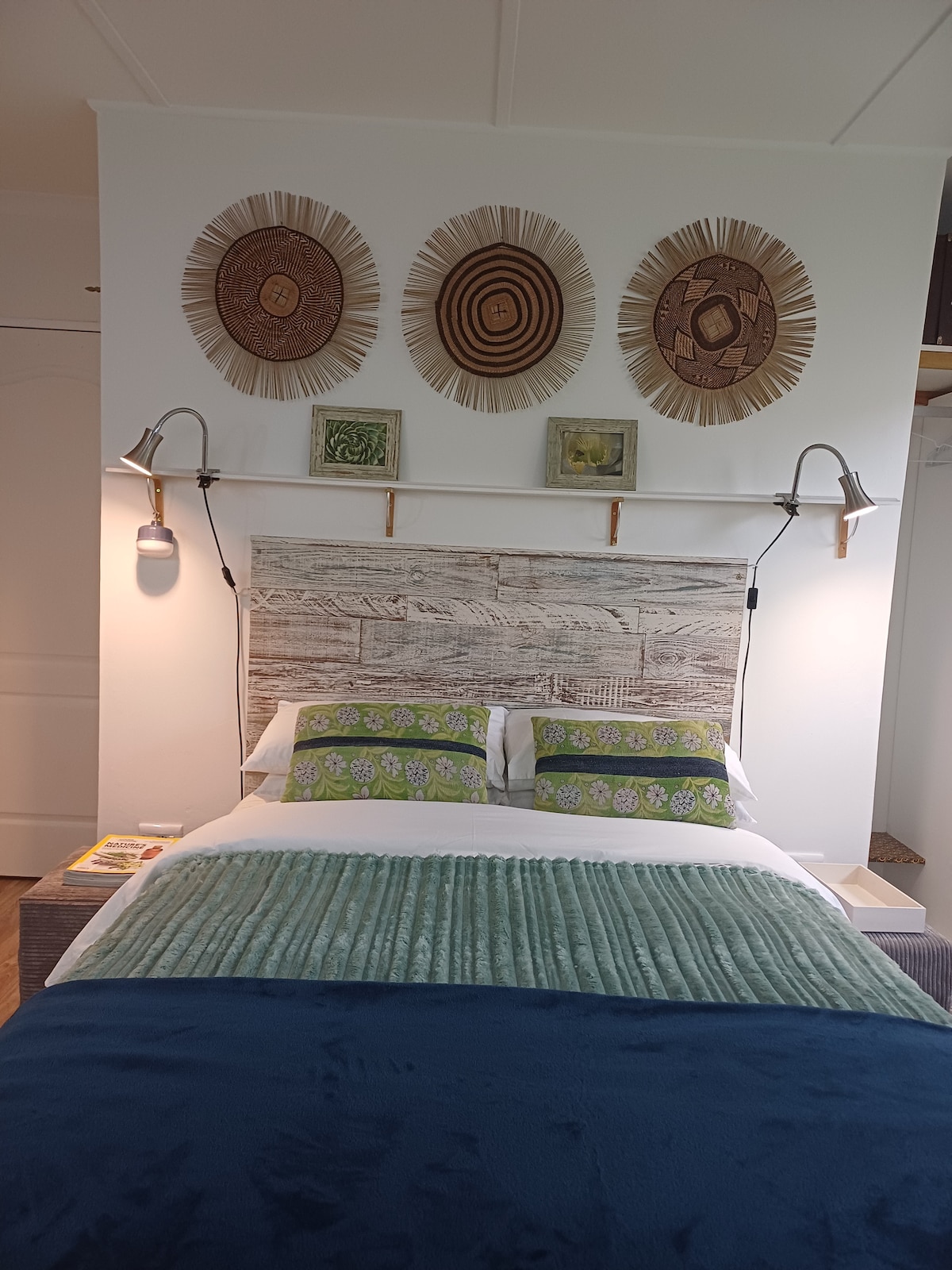 A cozy sleeping area features a spacious bed adorned with decorative green and floral pillows. Above the bed, three patterned wall hangings are displayed, complemented by framed artwork. Soft lighting is provided by wall sconces, enhancing the room's serene environment.