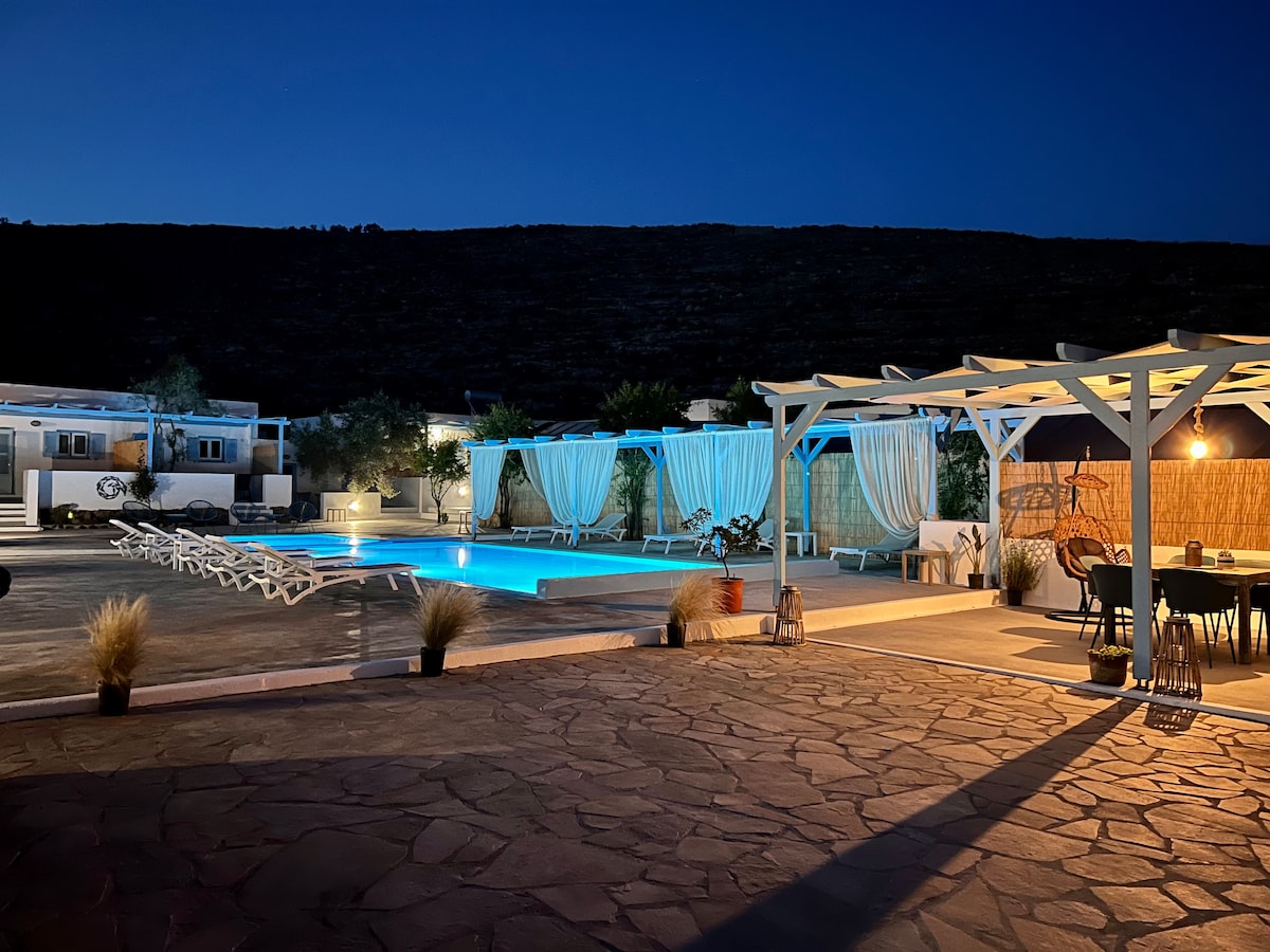 The outdoor area is highlighted by a sparkling swimming pool surrounded by sunbeds. Elegant cabanas with flowing curtains provide shaded seating. Soft lighting enhances the ambiance as evening approaches, and the surrounding landscape is gently illuminated, creating a serene atmosphere.