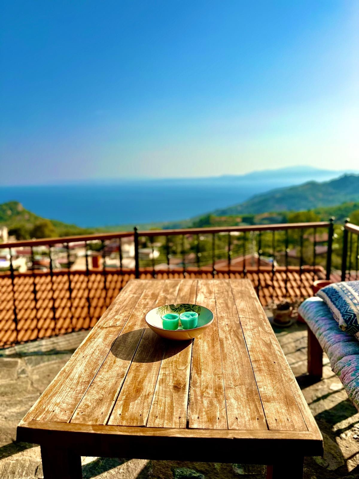 A wooden table is positioned outdoors, offering a clear view of the sea and surrounding landscape. A bowl with teal candles sits on the table, while a cozy blanket is draped over the side. The horizon is adorned with gentle hills under a bright sky.