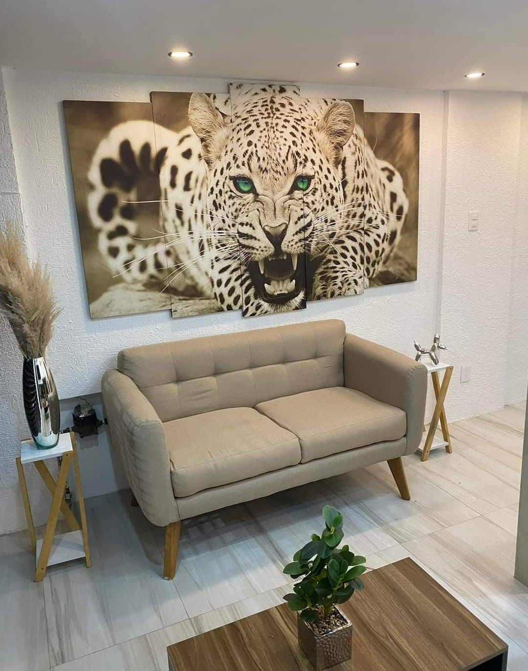 A beige sofa with a tufted design sits between two small side tables with decor. Behind it, a large artwork features a close-up of a white leopard, highlighting its striking green eyes. Indoor plants are positioned nearby, contributing to the room's ambiance.