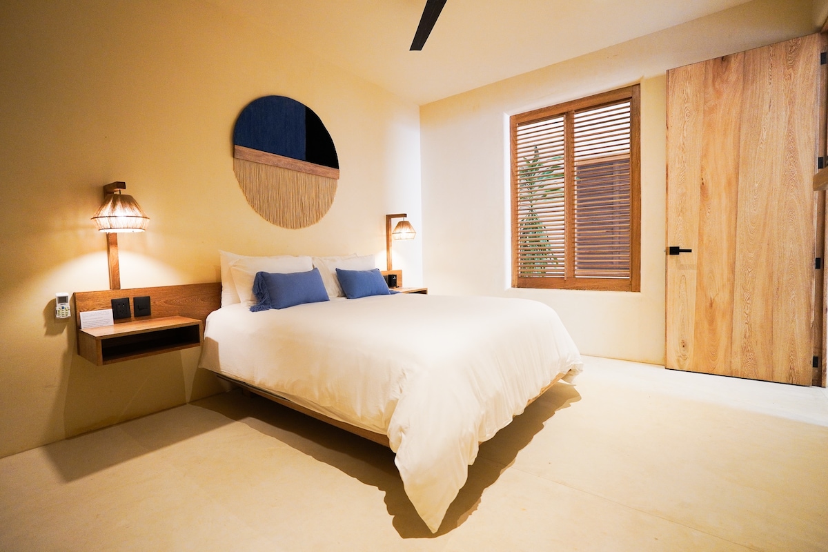 A serene bedroom features a spacious bed dressed in white linens, complemented by two pillows in soft blue. Simple wooden bedside tables house sculptural lamps, and natural light filters through a window with wooden blinds. A round mirror adds depth to the room's calm aesthetic.