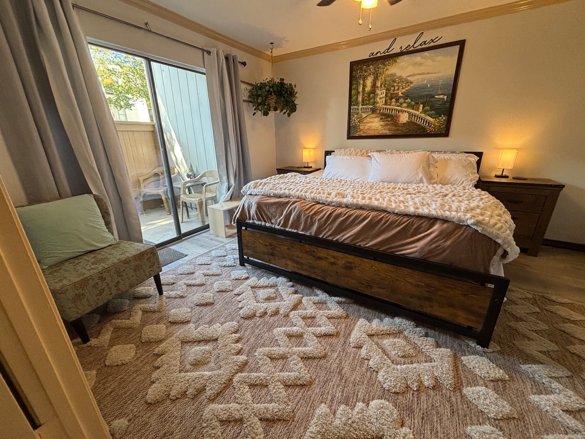 The bedroom features a spacious bed dressed in neutral linens, enhanced with a textured throw. Light filters through a sliding door leading to a private patio, while a small cozy chair and decorative artwork complete the warm atmosphere.