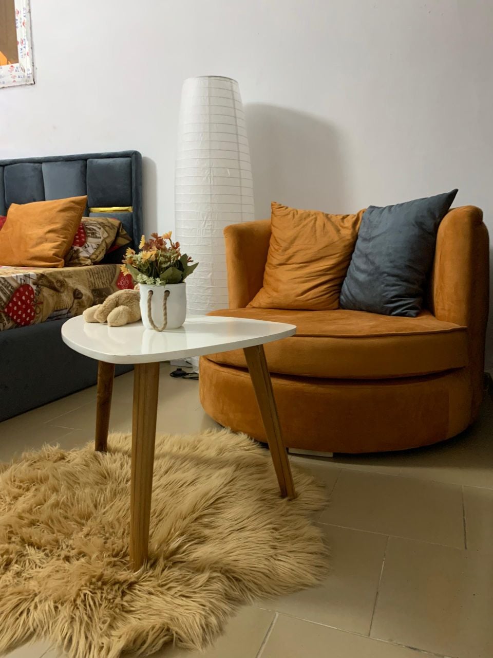 A cozy seating area features a round orange chair paired with blue and orange throw pillows. A small white table sits on a plush, light-colored rug. A decorative vase holds flowers, adding a touch of warmth to the space.