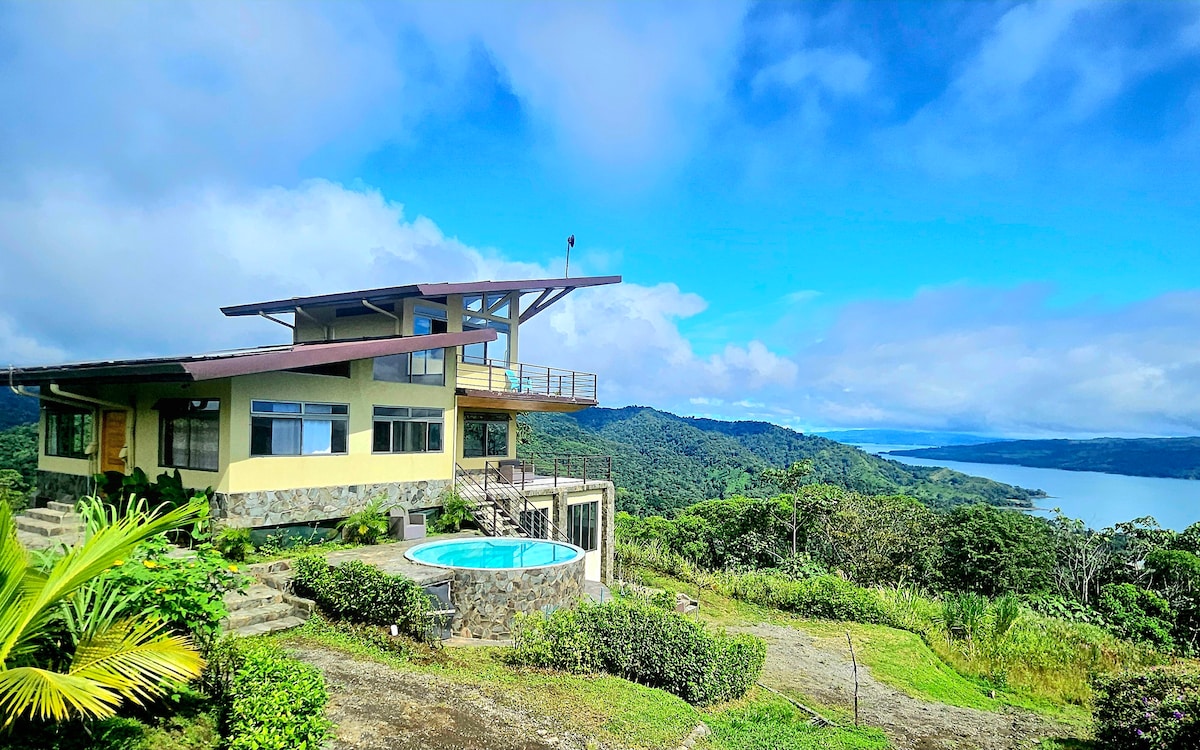 A modern retreat is set against a backdrop of lush mountains and a calm lake. The structure features a wrap-around balcony and large windows, offering expansive views of nature. A small lounge pool is visible in the foreground, surrounded by greenery.