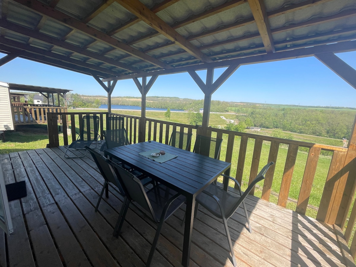 A covered outdoor deck features a large wooden table surrounded by six chairs. The area offers a scenic view of greenery and distant water, with light filtering through the open structure, creating a relaxed and inviting space for outdoor dining.