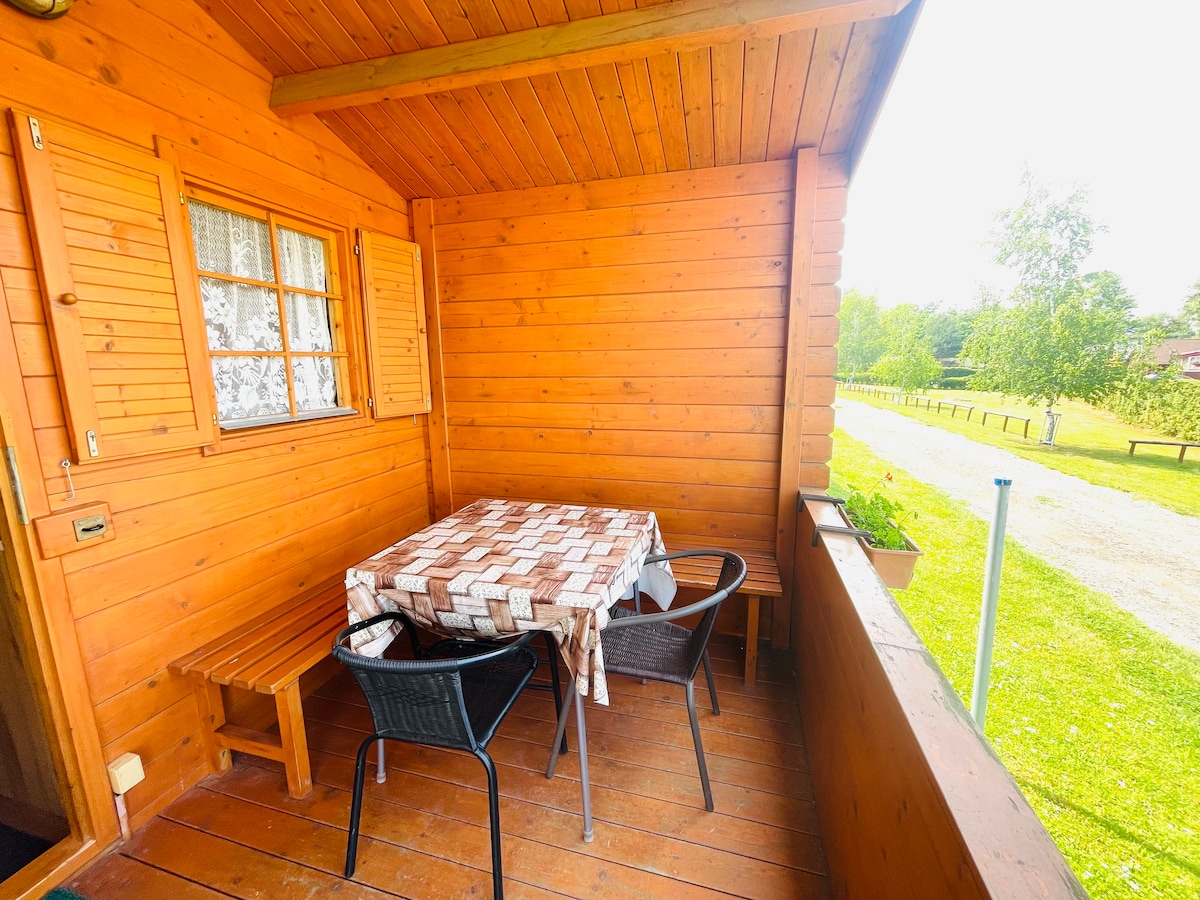A wooden balcony features a table covered with a checked cloth and four black chairs. A bench is positioned against the wall, and a scenic view of green fields and trees is visible beyond the railing, offering a peaceful outdoor space.