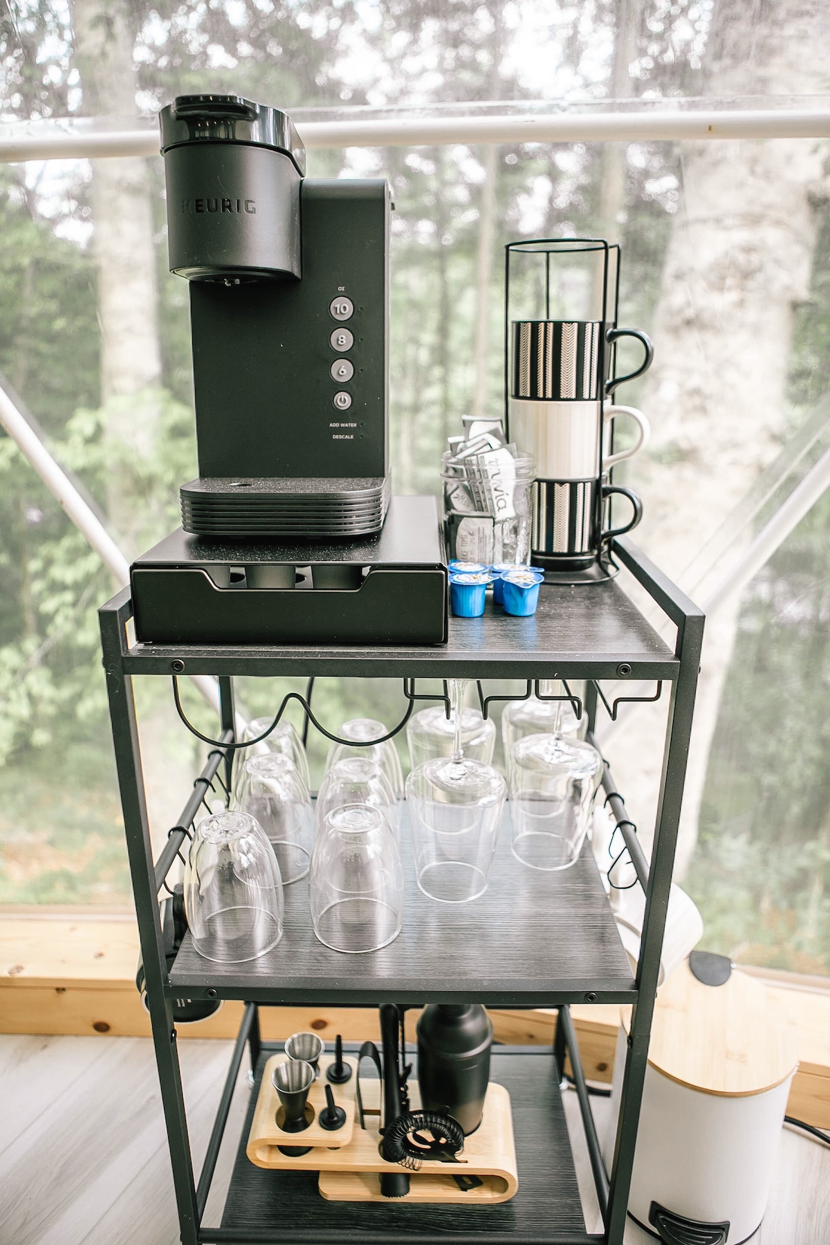 A sleek coffee station is presented, featuring a modern coffee maker with multiple brewing options. Below, an assortment of glasses and cups is organized on a two-tiered cart, complemented by additional coffee accessories, all set against a backdrop of large windows overlooking greenery.