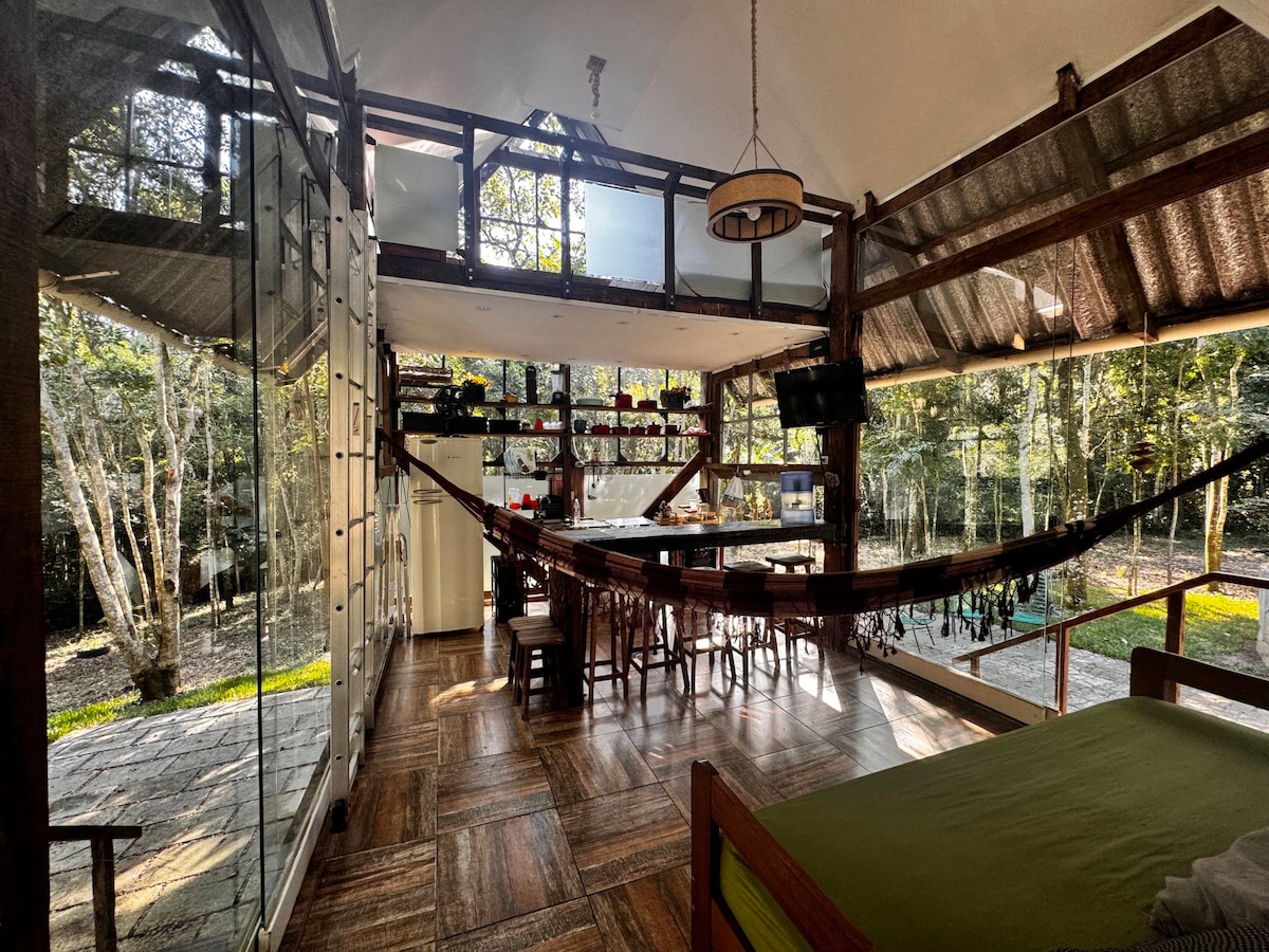 An open-concept interior showcases a blend of natural light and rustic decor. Wooden beams support the high ceiling, while a hammock is suspended between them. A kitchen area features shelves and a dining table, with large glass doors providing a view of the surrounding trees.