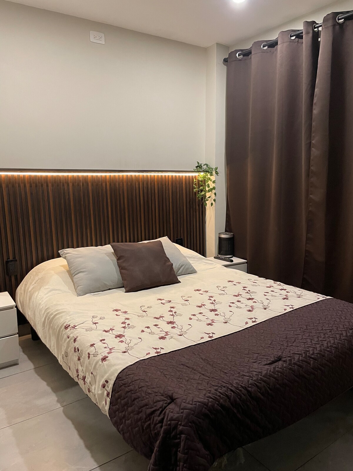 A serene sleeping area is presented with a spacious bed adorned with a floral-patterned blanket. Brown curtains frame the bed, and two nightstands are positioned on either side. Soft lighting outlines the headboard, complemented by a small plant for a touch of greenery.