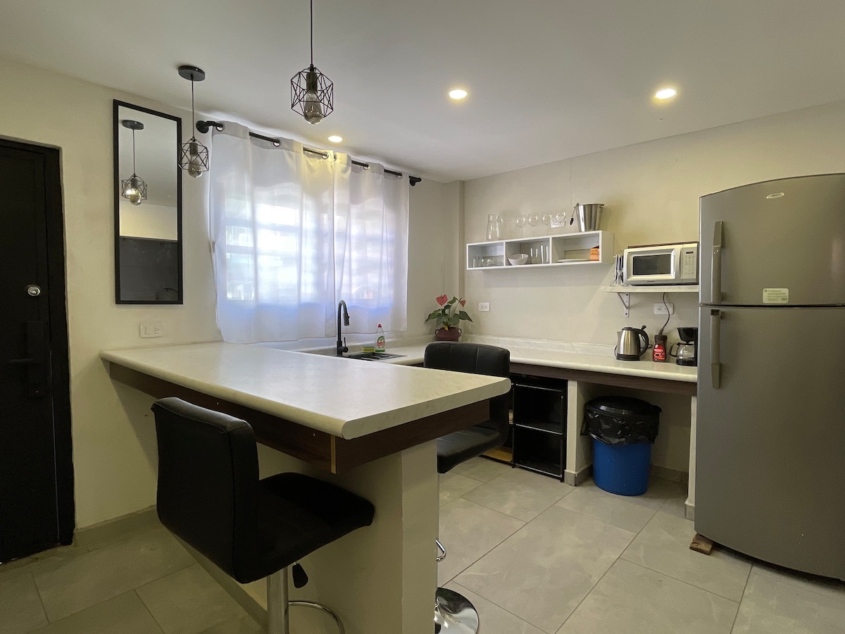 A modern kitchen features a long countertop with bar seating for two. Natural light filters through sheer curtains. A double stainless-steel sink and a variety of kitchen appliances, including a kettle, are visible. Neutral-colored cabinetry complements the tiled flooring.