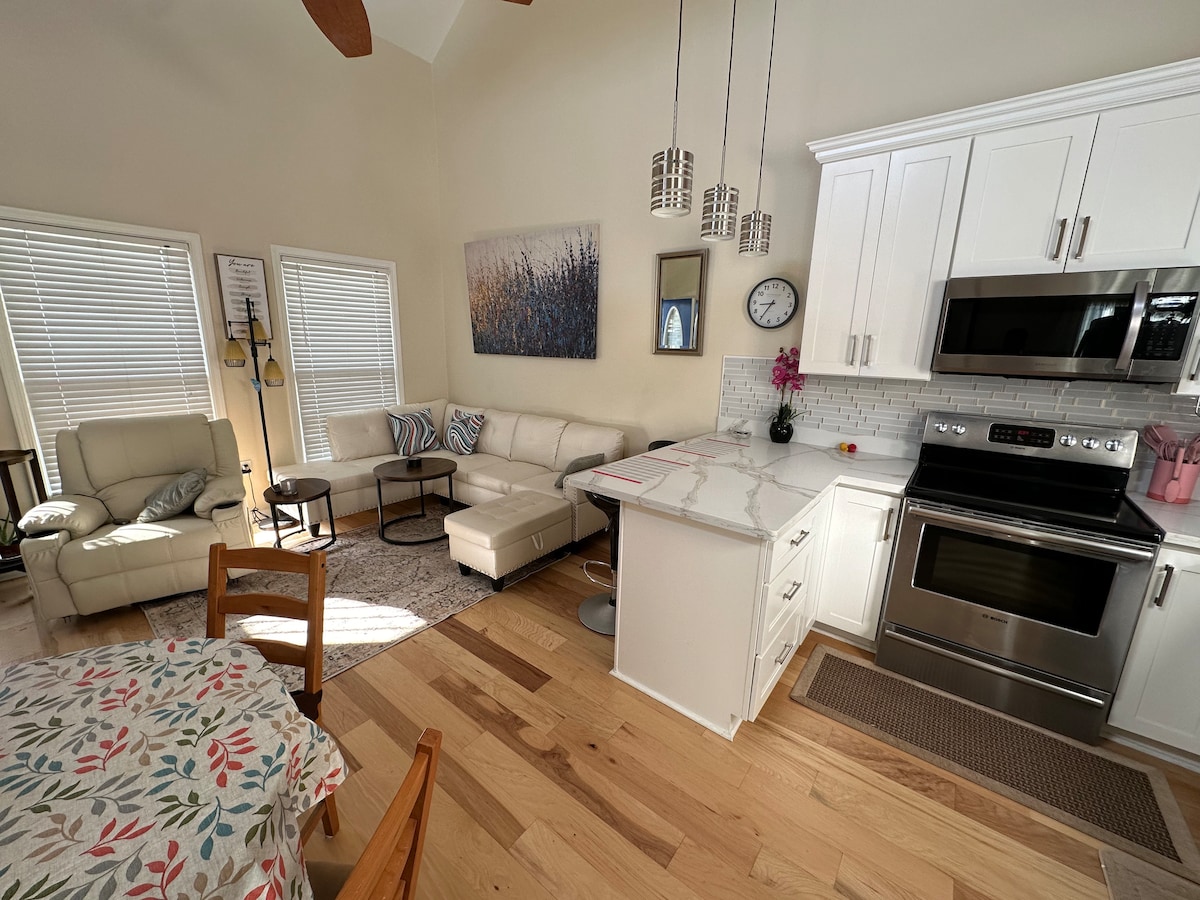 An open living area features a light-colored sectional sofa and a coffee table, with natural light from multiple windows. A kitchen island with bar seating is positioned near stainless steel appliances. A dining table with a floral tablecloth completes the inviting space.