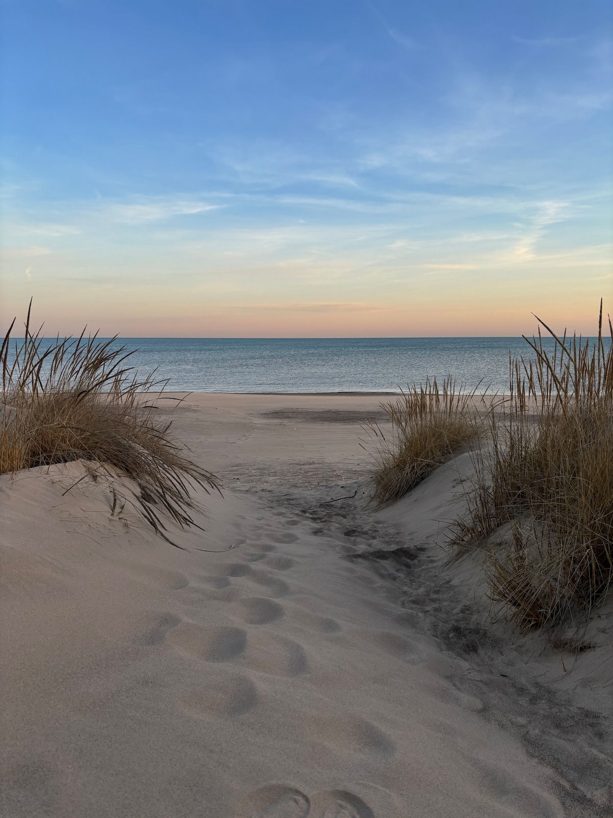 A serene beach scene is depicted, with gentle sand dunes framed by tall grasses. Footprints lead toward the calm, blue sea, where the horizon merges with a pastel sky at dusk. The tranquil environment invites relaxation and exploration.