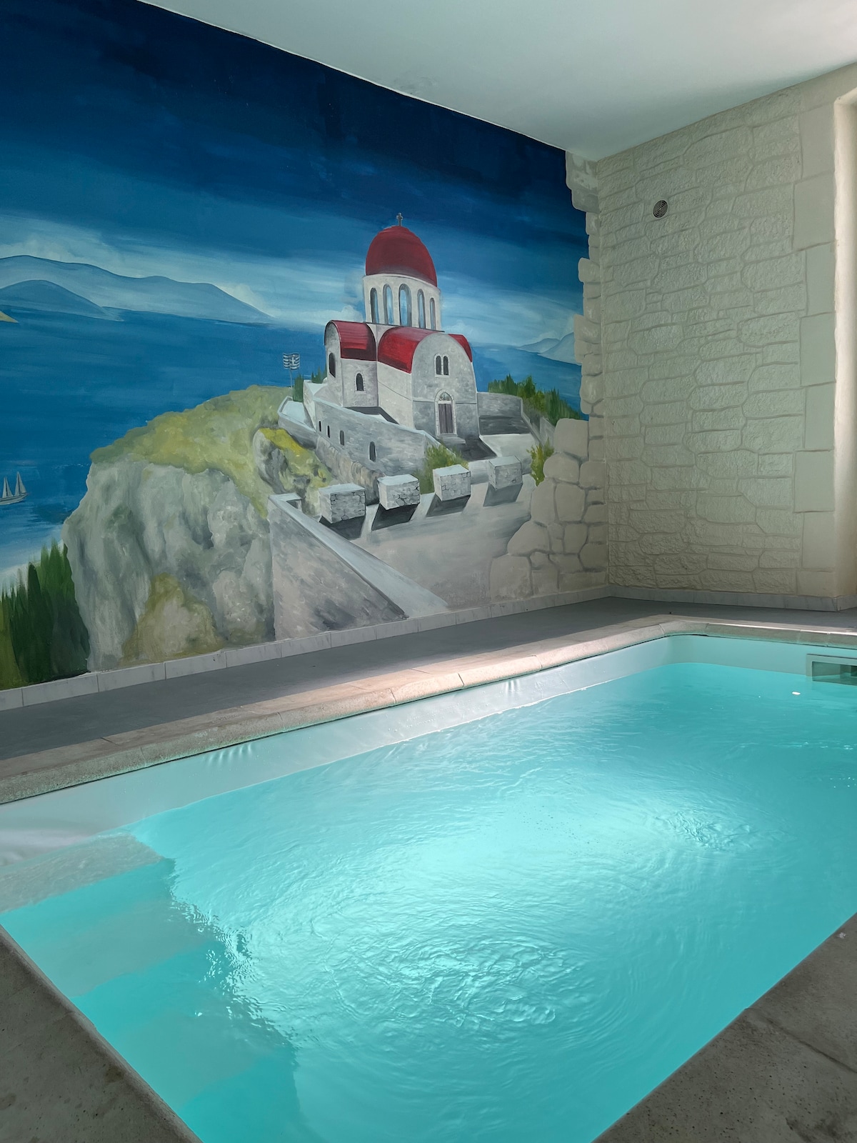 An indoor pool is set against a colorful mural depicting a coastal landscape with a red-domed church. The pool features clear, inviting water, surrounded by light-colored stone walls that enhance the spacious feel of the area.