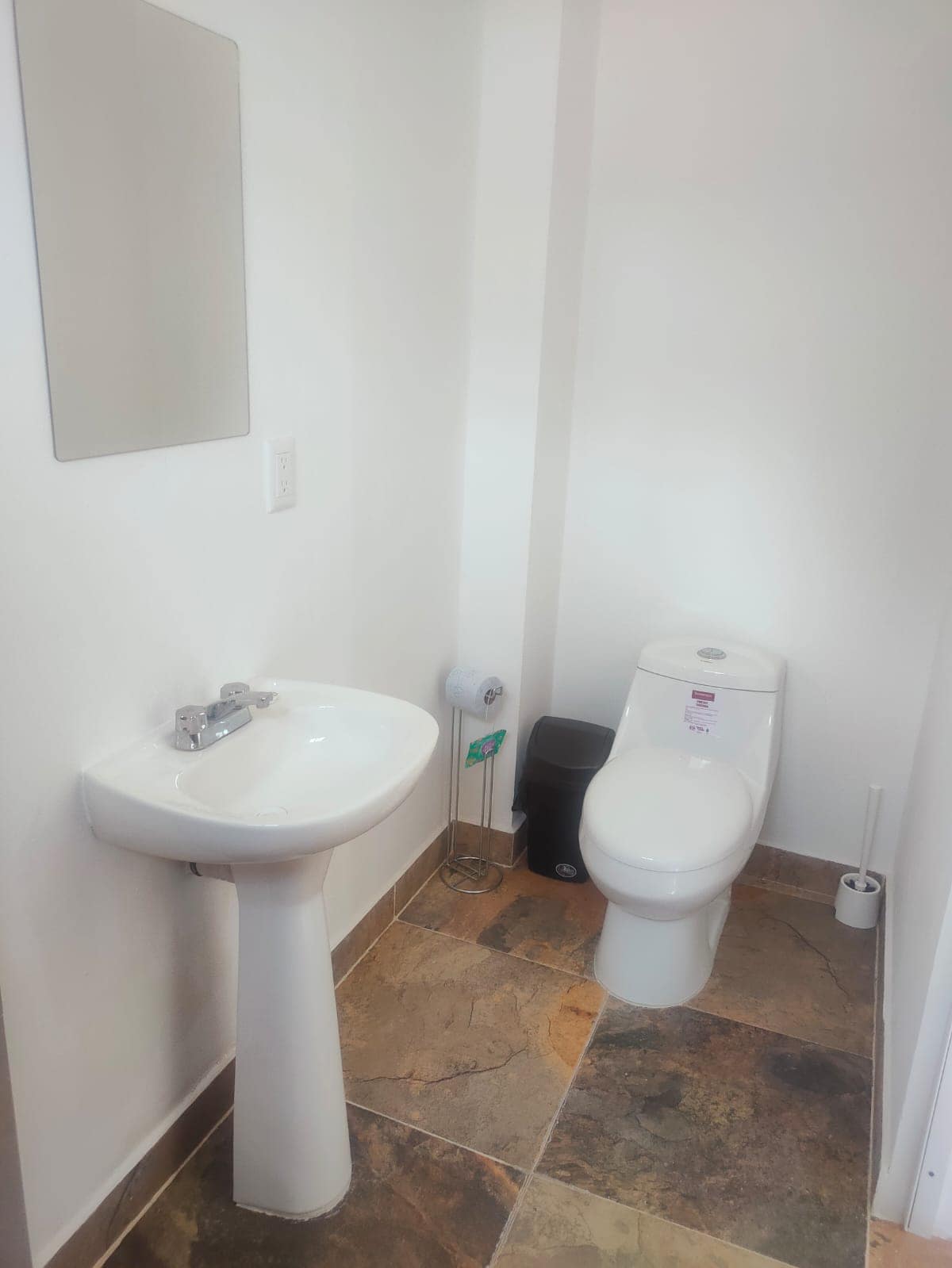 A clean and modern bathroom features a compact toilet and a pedestal sink with a single faucet. Sleek tiles in earthy tones cover the floor, while natural light illuminates the space. A small waste bin is also visible.