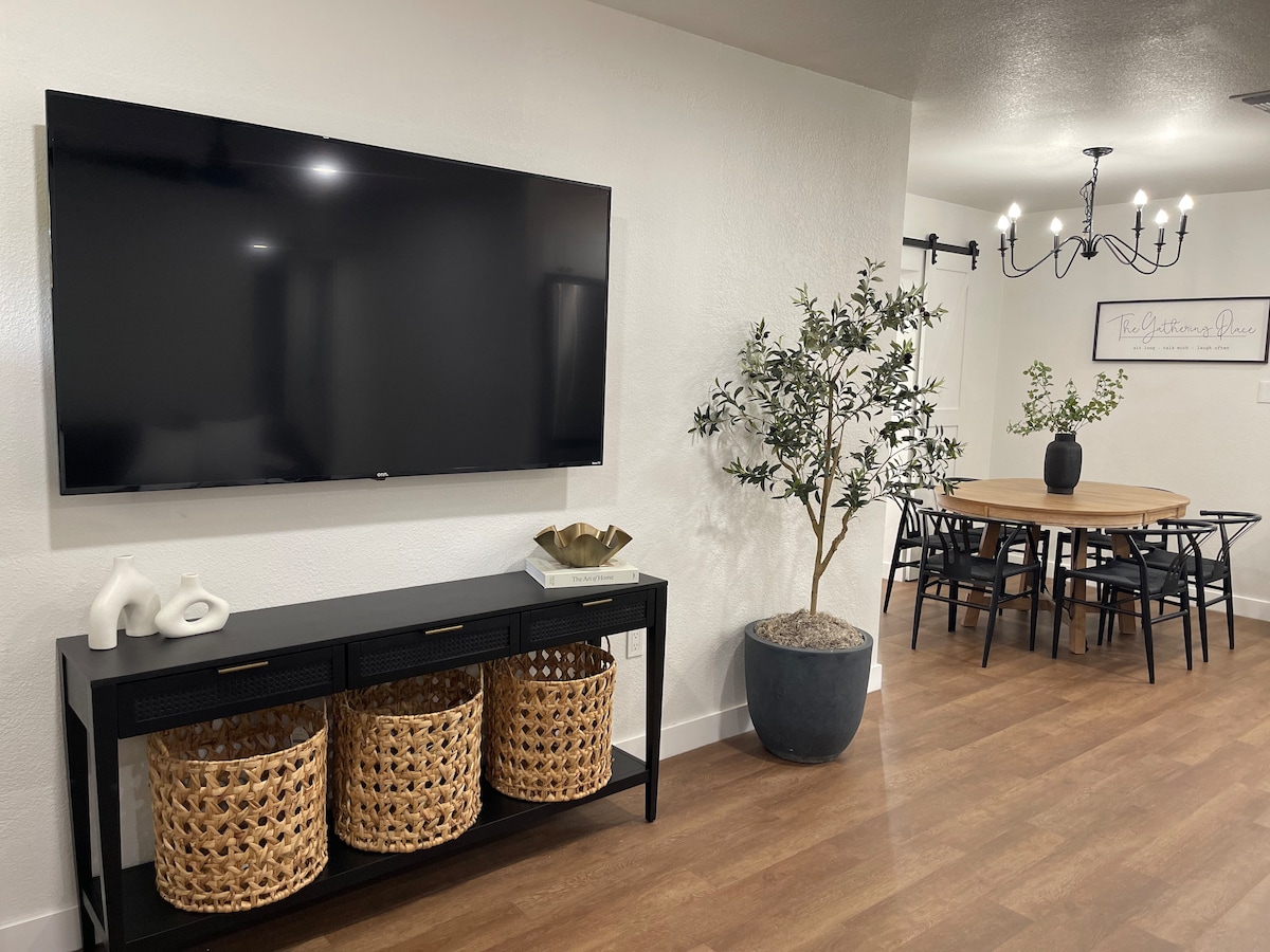 A living area features a mounted flat-screen TV above a console table with woven baskets below. A dining area is visible in the background, showcasing a round table surrounded by black chairs and a decorative plant in a large pot.