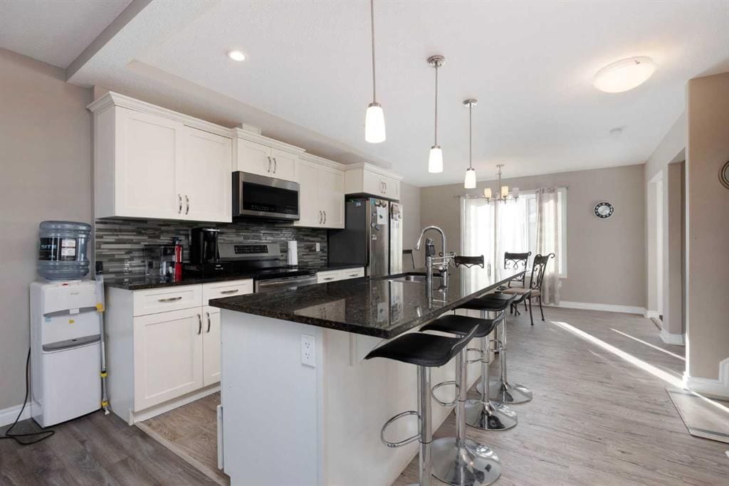 A modern kitchen features light-colored cabinetry and dark granite countertops. Stainless steel appliances include a refrigerator and stove. Ample natural light enters through large windows, illuminating the bar area with four sleek stools. A dining area with a table and chairs sits in the background.