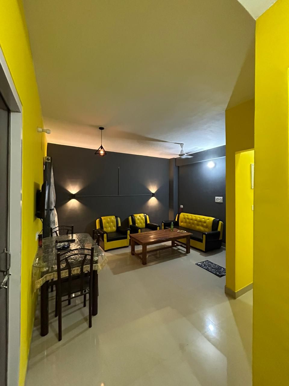 The living area features stylish yellow and gray walls, complemented by modern seating arrangements. A wooden coffee table occupies the center, with overhead lighting casting a warm glow. A dining table is positioned near the entrance, creating a cozy and inviting environment.