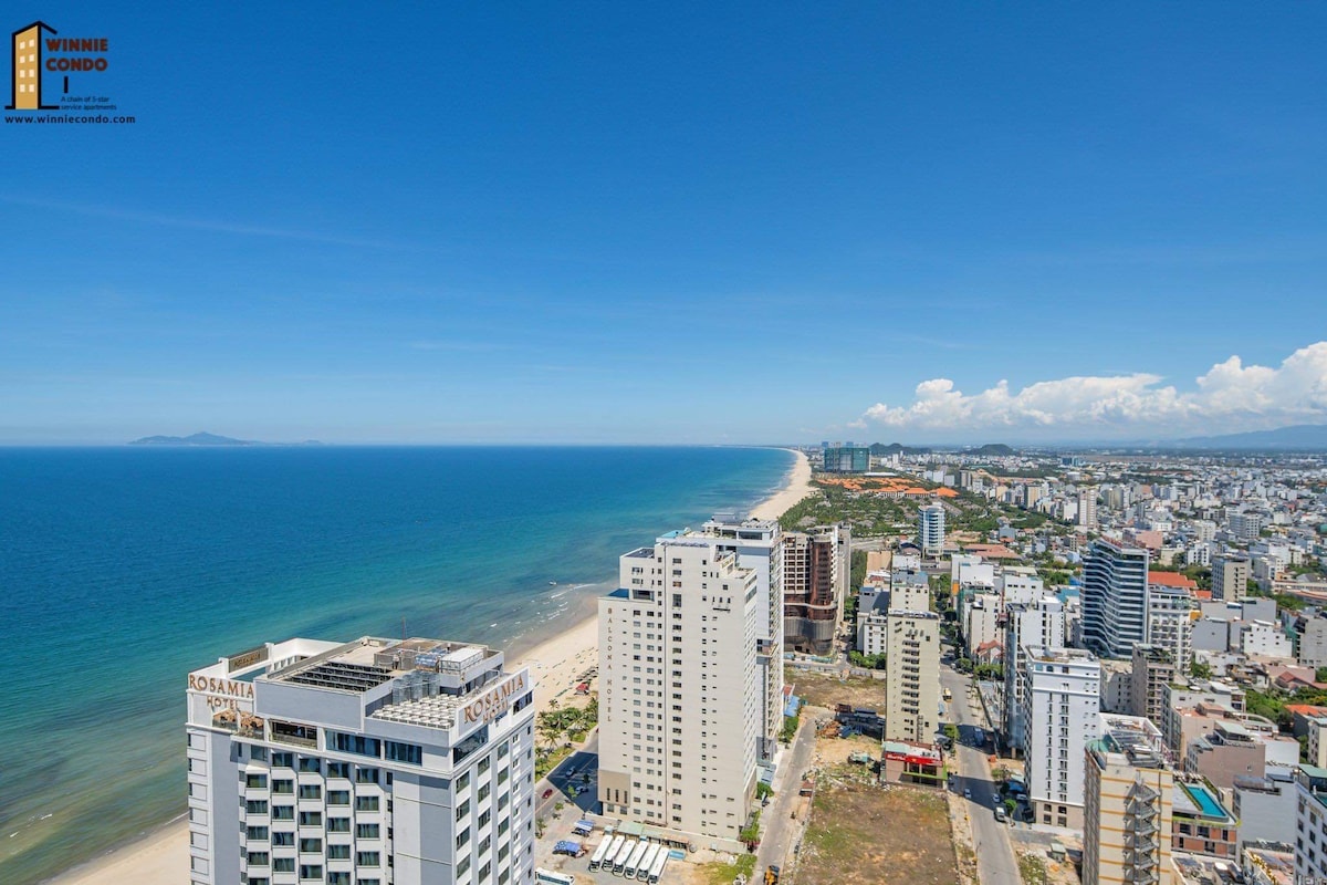 An expansive view of the coastline highlights the golden beach stretching along the vibrant blue ocean. The skyline is adorned with a mix of modern buildings and lush green mountains in the distance, with clear skies above creating a serene backdrop.