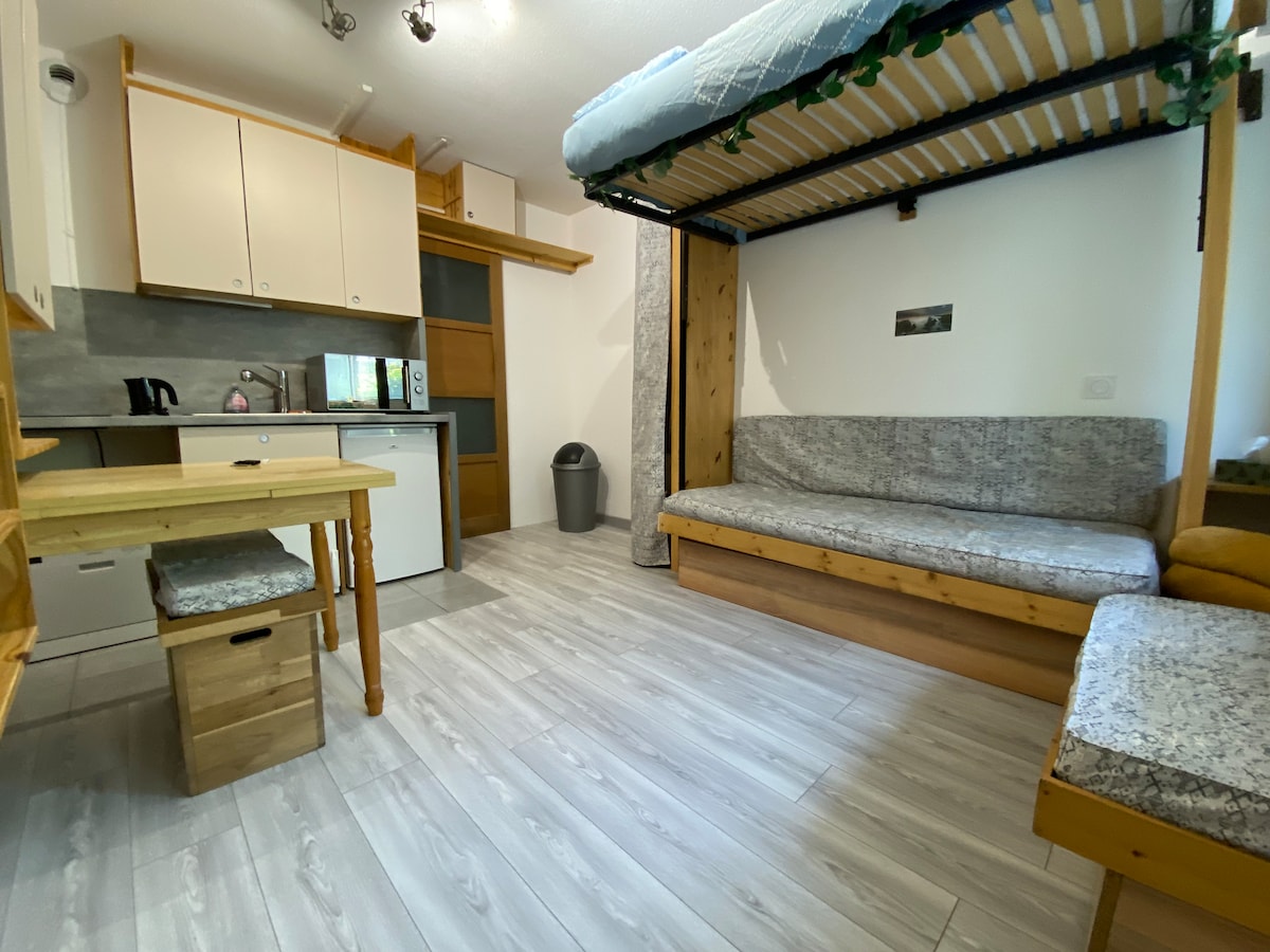 A studio space featuring a wooden lofted bed above a comfortable sofa area. A small dining table and two stools are positioned near the kitchenette, which includes essential appliances. The light wood tones and neutral colors create a calming atmosphere, with sliding doors allowing natural light.