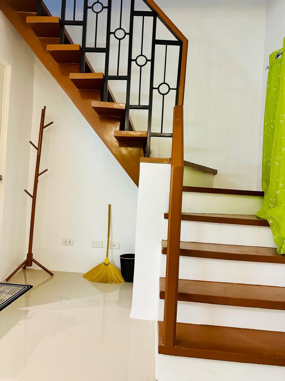 The image showcases a two-tone staircase featuring wooden steps and a decorative metal railing. A sleek coat rack is positioned nearby, along with a broom and a small wastebasket. A bright green curtain adds a pop of color to the space.
