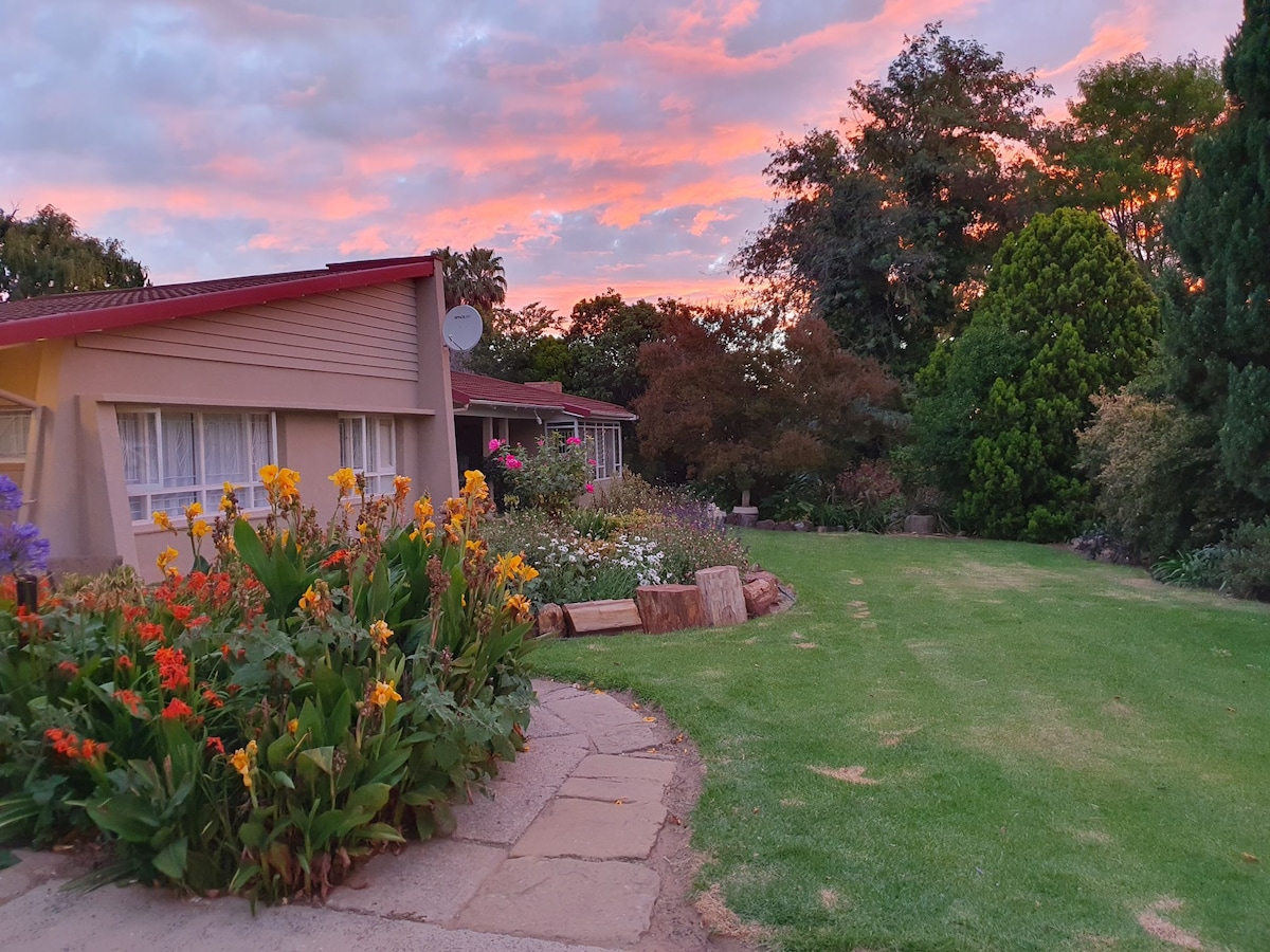 A tranquil outdoor area features a well-maintained garden filled with colorful flowers, set against a vibrant sunset sky. The neatly trimmed grass leads to a cozy home with large windows, providing a peaceful retreat in a quiet environment.