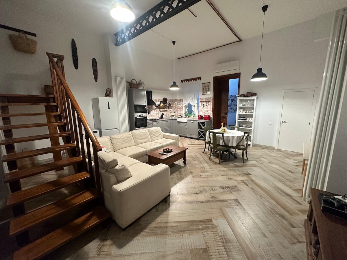 An open-plan living area features a spacious beige sofa and a wooden coffee table. A dining table is set for four, and modern kitchen appliances are visible. Natural light floods the space through a large window, while a staircase leads to an upper level.