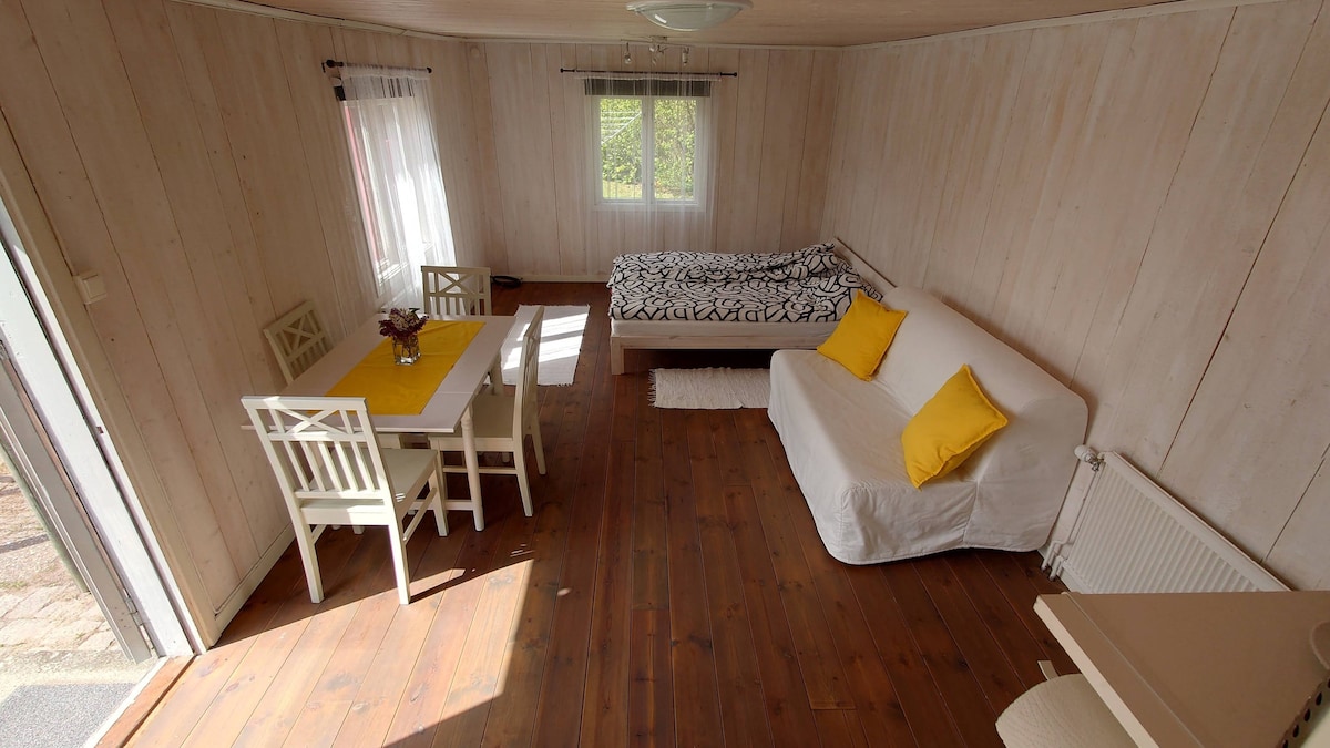 The interior of the tiny wooden house is arranged with light wooden walls and a warm wooden floor. A dining table is set with a yellow tablecloth, accompanied by four white chairs. A sleeping area features a double bed and a sofa, both adorned with yellow cushions.