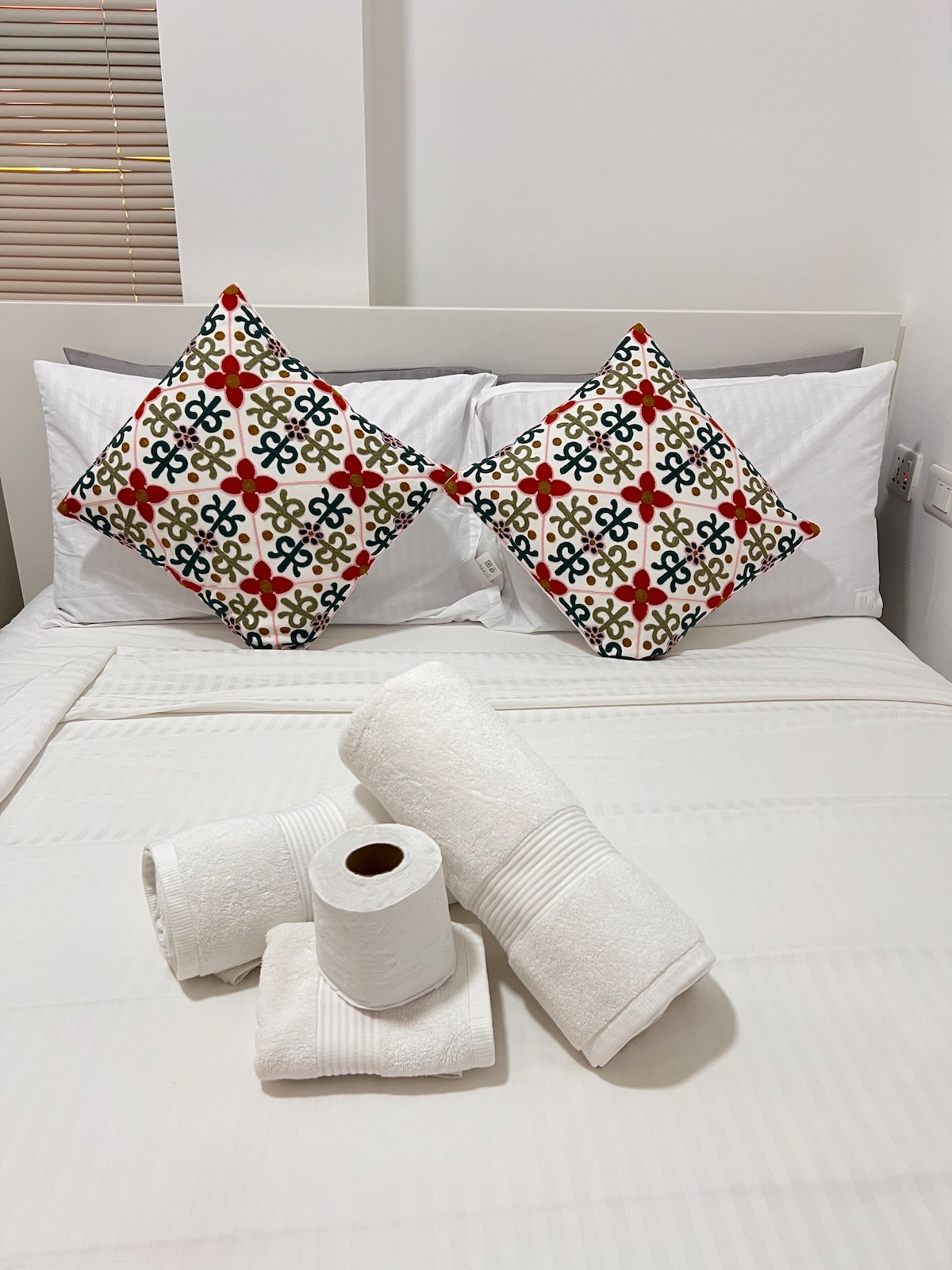 A bedding area features a spacious bed with crisp white linens. Colorful decorative pillows are arranged on the headboard. Rolled towels and toilet paper are neatly placed at the foot of the bed, contributing to a fresh and welcoming ambiance.