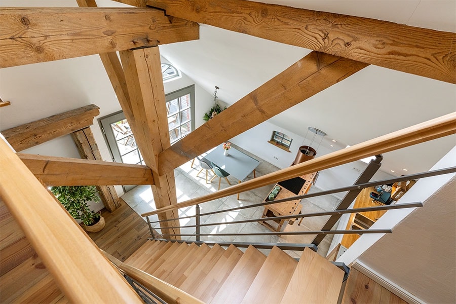 A view from above captures the interior's open design with exposed wooden beams and a staircase leading down. Natural light filters through windows, illuminating the dining area and furnishings below, while a potted plant adds a touch of greenery.