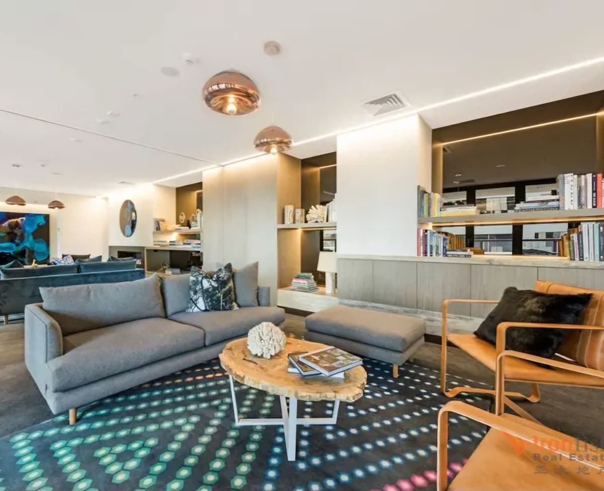 A modern living area features a neutral-toned sofa and a wooden coffee table, complemented by a patterned rug. Recessed lighting and decorative lamps illuminate the space, while shelving displays various books and decor items in the background.