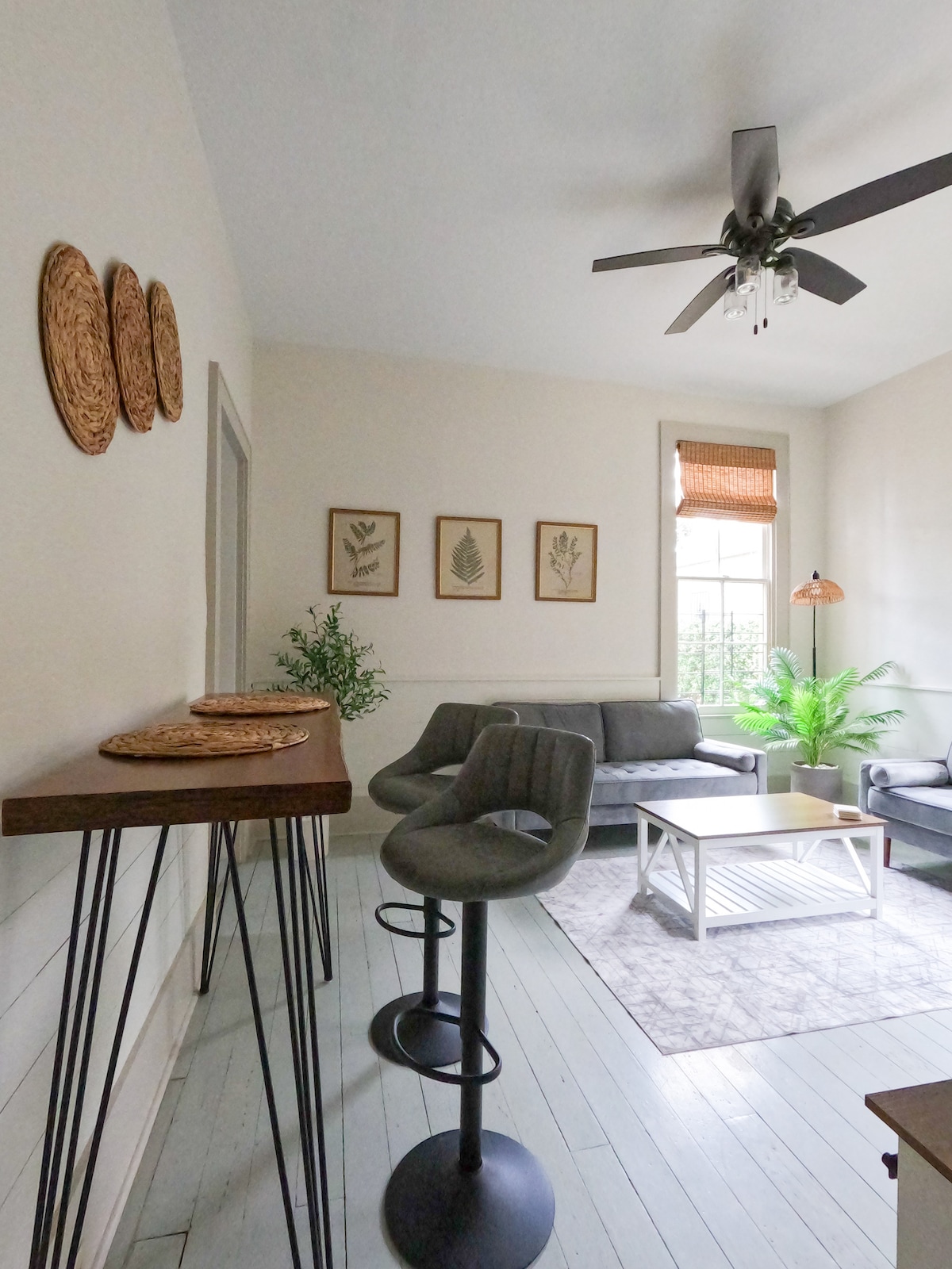 A cozy living area is shown, featuring a light-colored rug and a coffee table surrounded by plush gray seating. Wall art showcasing botanical prints is displayed above a console table with woven placemats. A ceiling fan and natural light from the window add to the ambiance.