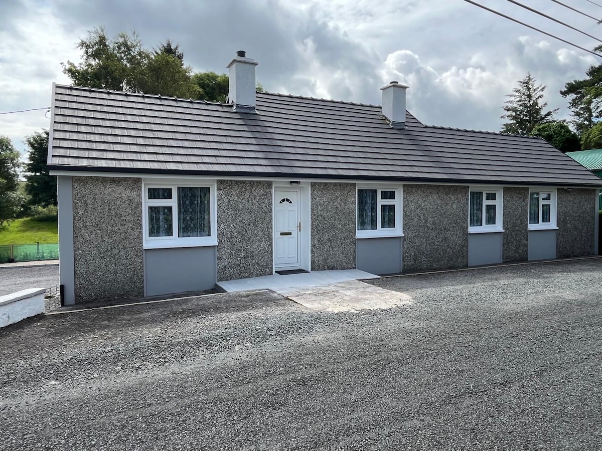 A single-story cottage is depicted, with a textured exterior and a pitched roof. The front entrance features a central door flanked by two windows. Lush greenery is visible in the background, with a gravel driveway leading up to the entrance.