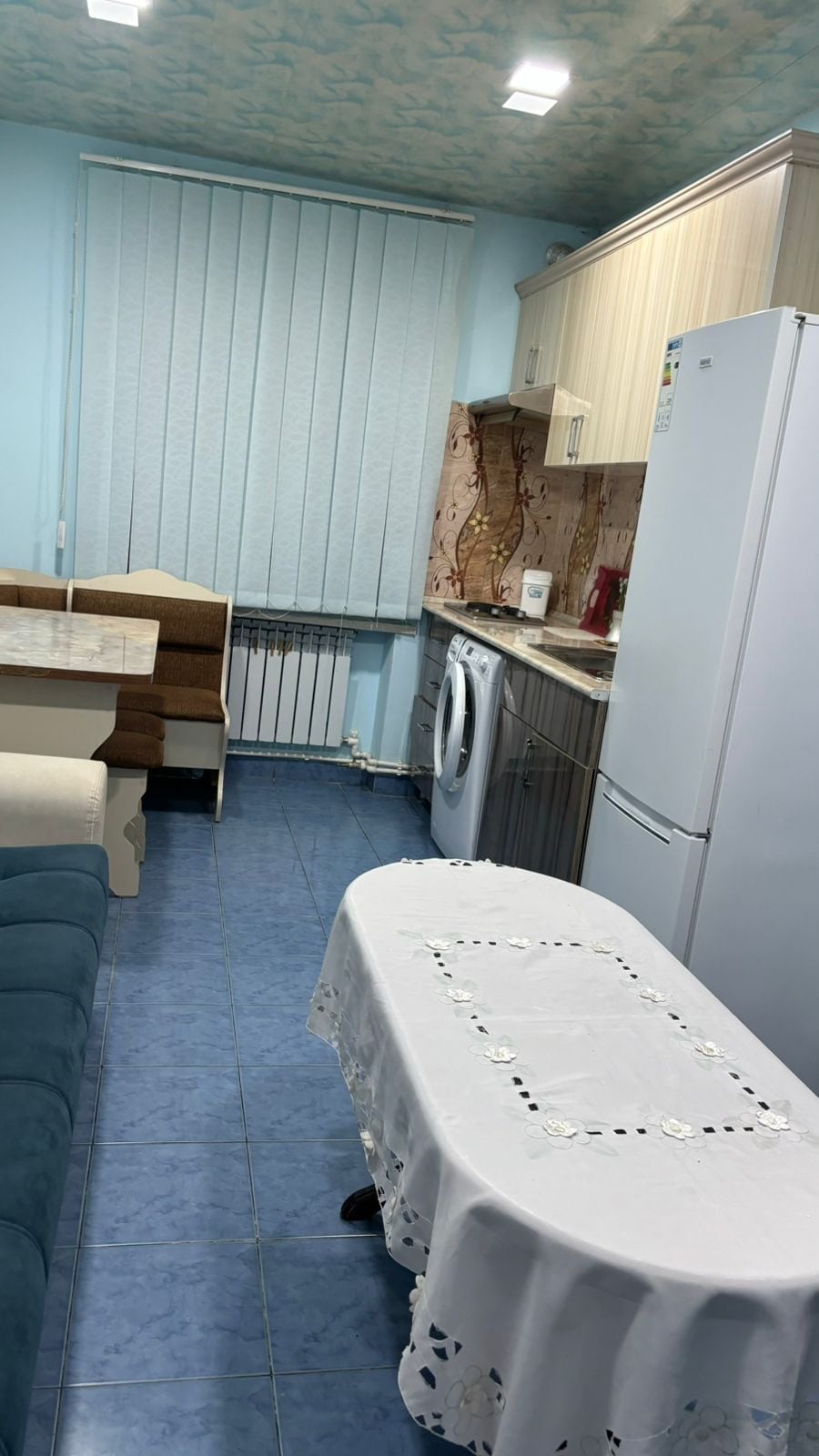 A fully equipped kitchen area features light cabinetry and modern appliances, including a refrigerator and washing machine. A dining table is set with a white tablecloth and surrounded by cushioned seating. Natural light enters through vertical blinds covering a window.