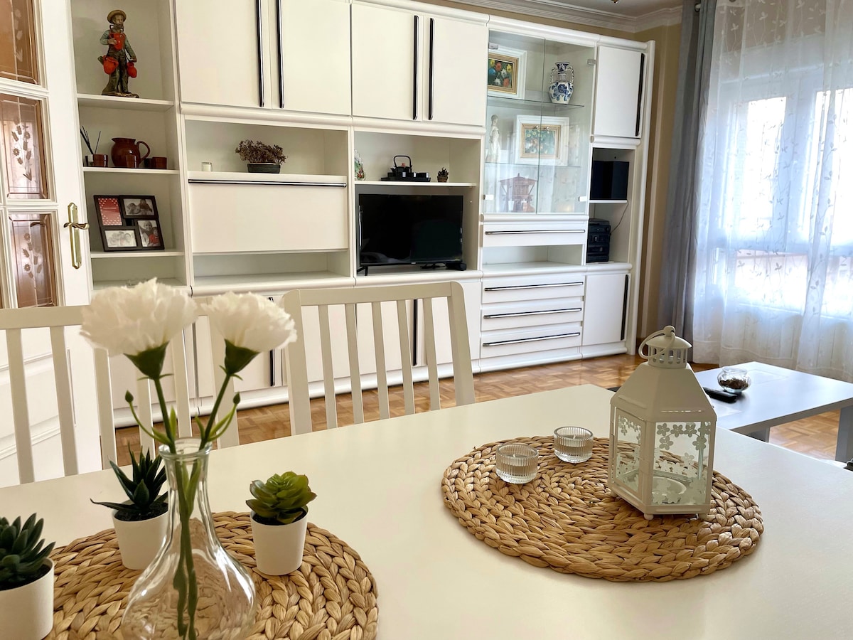 A dining area is featured, showcasing a white table surrounded by modern chairs. A decorative woven placemat holds small potted plants. A lantern and candles add a subtle touch beside a vase with white flowers. Natural light filters in through large windows.