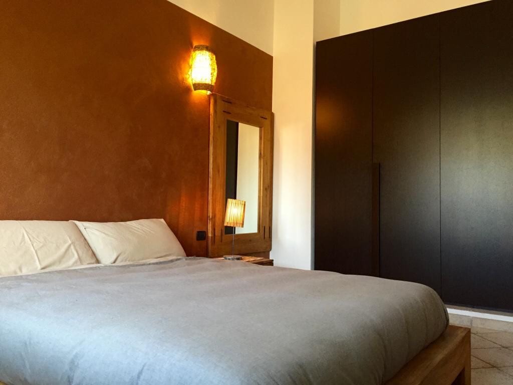 A spacious bedroom is highlighted by warm, earthy tones on the walls, with a large bed featuring neutral bedding. A wooden mirror is positioned above a nightstand beside the bed, and a black wardrobe stands against the wall, contributing to the room's modern aesthetic.