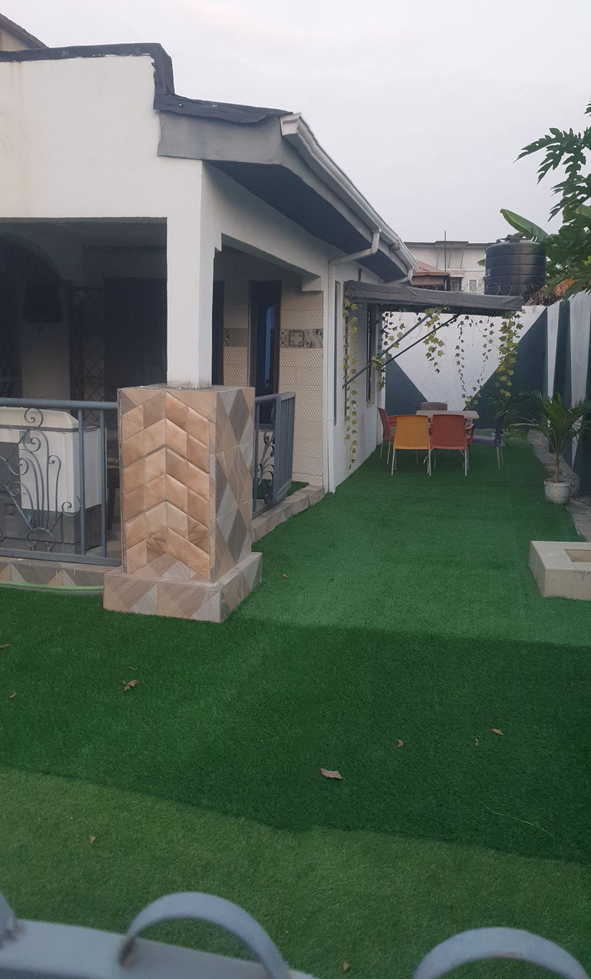 A neatly landscaped area features artificial grass and a small patio set with colorful chairs. The exterior of the bungalow is visible, complemented by decorative greenery and a charming stone wall that enhances the inviting atmosphere.