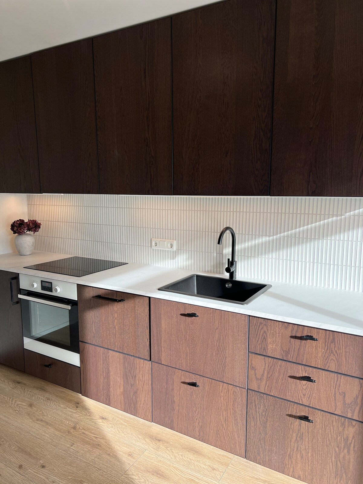 A modern kitchen is shown, featuring dark wooden cabinetry and a light-colored countertop. A single basin sink and a black faucet are positioned next to an oven and cooktop. Decorative items are placed on the countertop, adding a subtle touch of elegance.