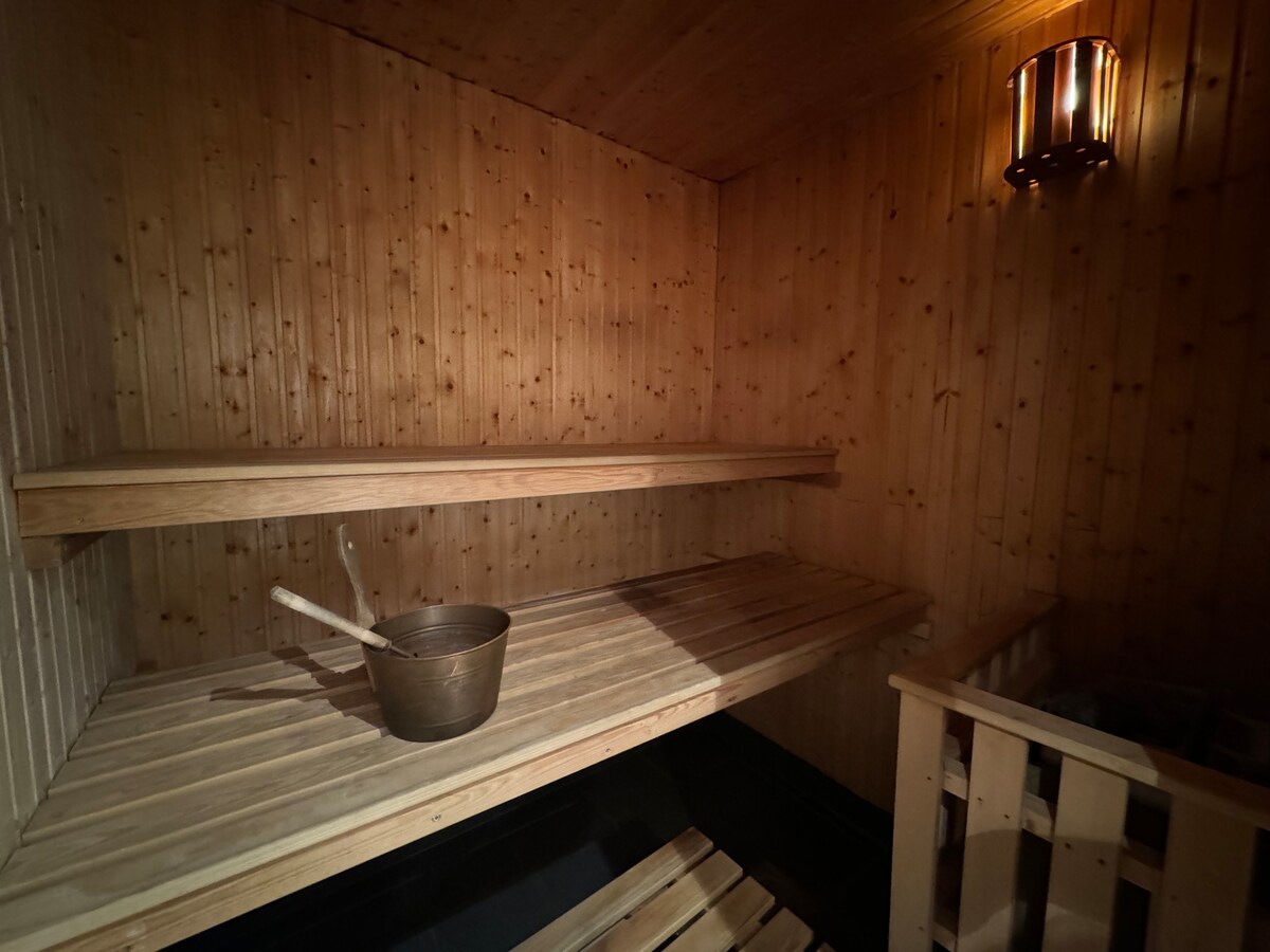 A wooden sauna is presented, featuring two wooden benches along the walls. A metal bucket and ladle are placed on one of the benches, and a small wooden structure, likely for personal items, is visible. Soft lighting enhances the warm wooden interior.
