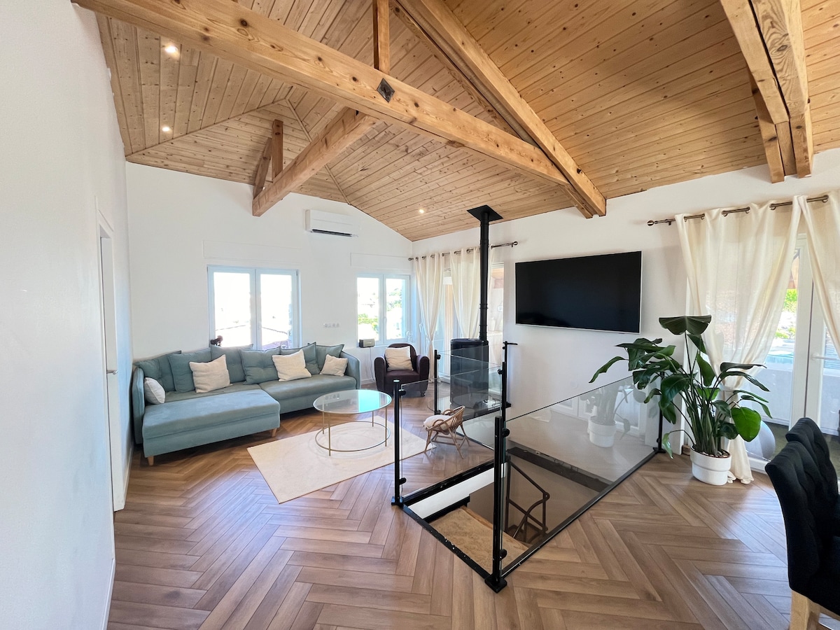 A bright and airy living space features a combination of wooden beams and large windows. A comfortable sofa is positioned near a glass coffee table, while a potted plant adds a touch of greenery. A television is mounted on the wall, providing a focal point in the room.